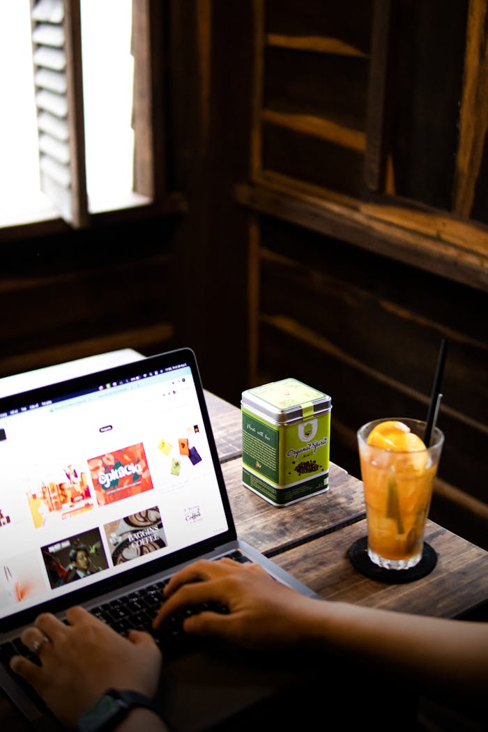 Close-up of hands typing on a laptop beside a chilled drink at a wooden table in a cozy setting.